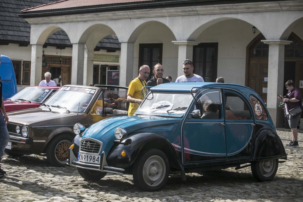 Na płycie rynku Miasteczka Galicyjskiego stoją zabytkowe samochody w różnych kolorach. Przy autach znajdują się ludzie i oglądają pojazdy.  