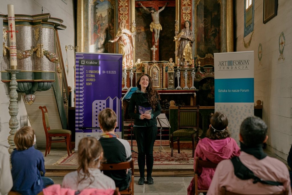 Kobieta zapowiada artystów podczas Festiwalu Akordeonowego w kościele rzymskokatolickim w Sądeckim Parku Etnograficznym. Stoi pomiędzy dwoma banerami, które reklamują koncert. Przed kobietą siedzi pięć osób. 