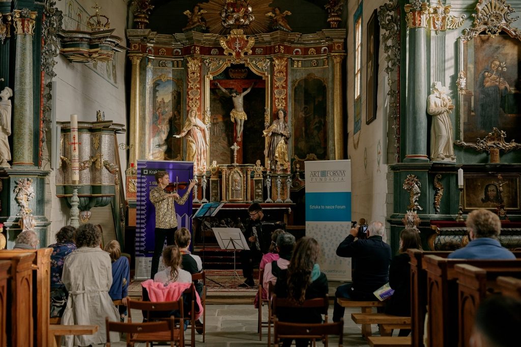 Kilkanaście osób słucha koncertu koncert muzyki akordeonowej w kościele rzymskokatolickim w Parku Etnograficznym w Nowym Sączu. Gra akordeonista oraz skrzypek. 