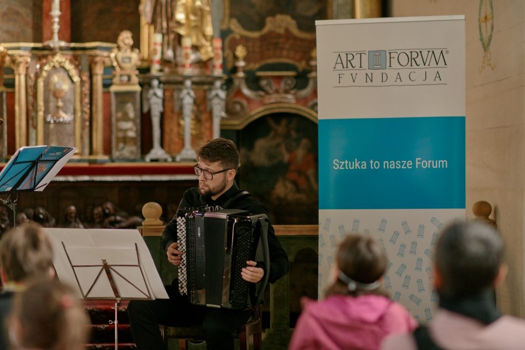 Kilka osób słucha koncertu akordeonowego w kościele rzymskokatolickim w Parku Etnograficznym w Nowym Sączu. Gra jeden akordeonista.