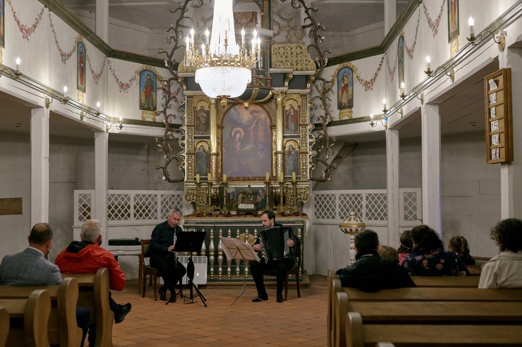 Dwóch mężczyzn gra na instrumentach. Na nimi ołtarz. Słucha ich kilka ludzi. 